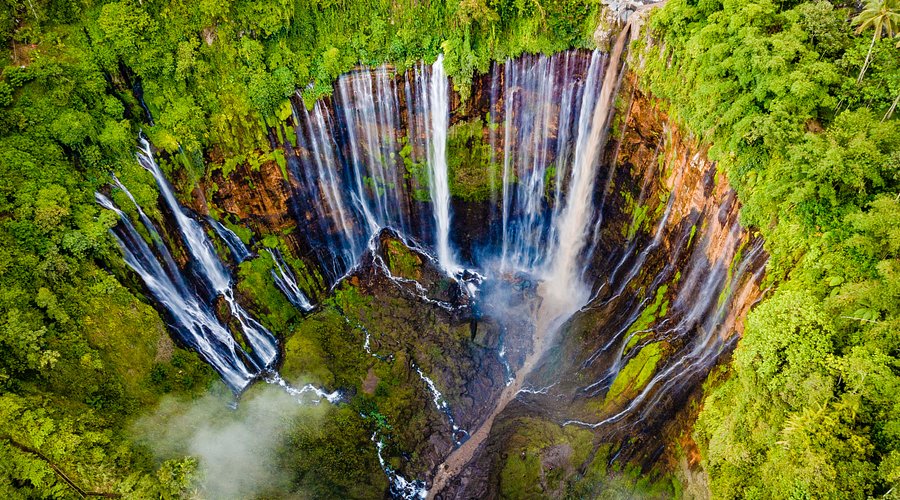 [An aerial view captures the 'thousand waterfalls' effect of Tumpak Sewu, cascading down a lush, green cliffside in East Java.]