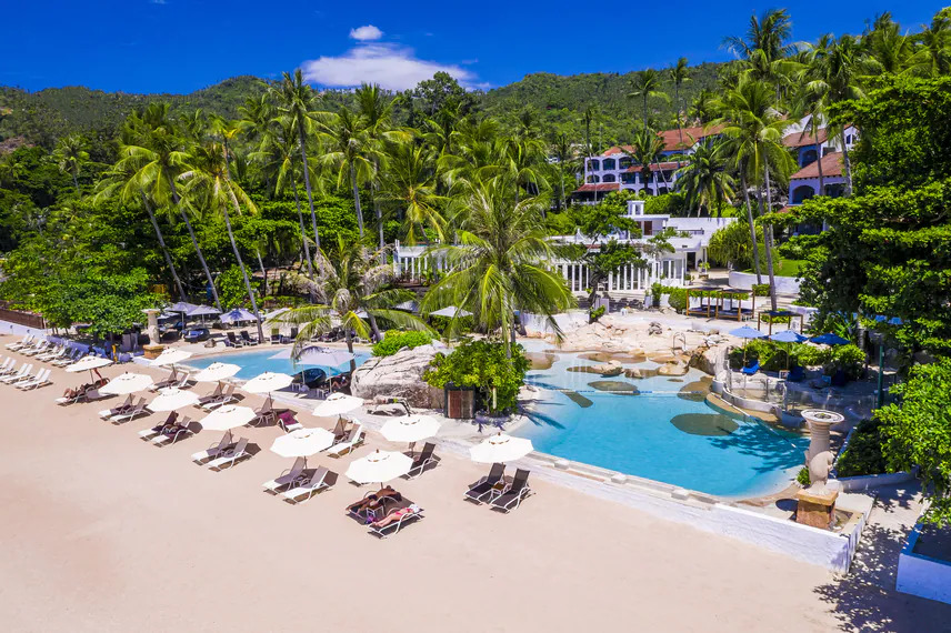 An aerial view of the stunning lagoon-style swimming pool at the Sheraton Samui Resort, nestled amongst palm trees right by the beach.