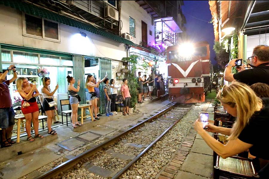 [A crowd of locals and tourists gather to watch a train pass through the narrow residential street in Hanoi.]